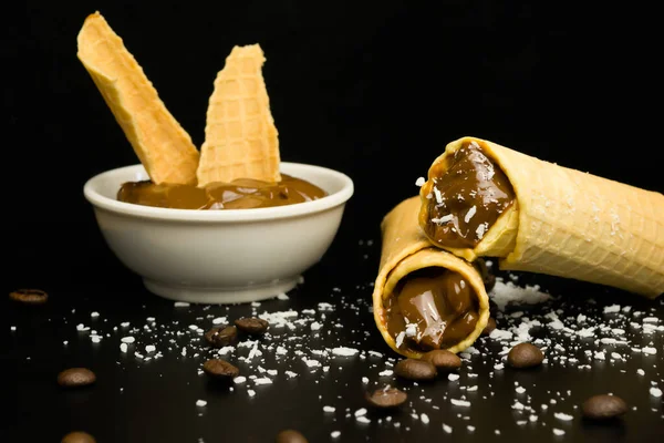 Wafer rolls with condensed milk and a saucer in which there is a filler for wafers on a black background