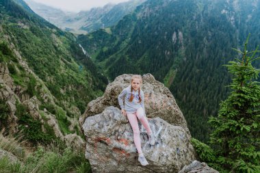 Güzel bir kız dağlarda bir kayanın üzerinde oturur ve bir dağ vadisinin güzel manzarasına bakar..