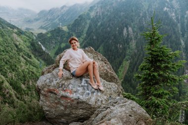 Güzel bir kız dağlarda bir kayanın üzerinde oturur ve bir dağ vadisinin güzel manzarasına bakar.