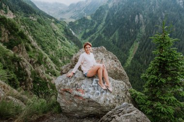 Güzel bir kız dağlarda bir kayanın üzerinde oturur ve bir dağ vadisinin güzel manzarasına bakar.