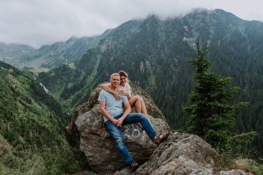 Çekici bir kadın ve kocası arka planda dağlarla birlikte kayaların üzerinde oturuyor. 