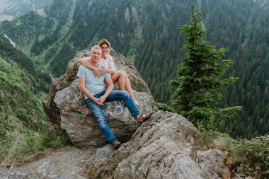 Çekici bir kadın ve kocası arka planda dağlarla birlikte kayaların üzerinde oturuyor. 