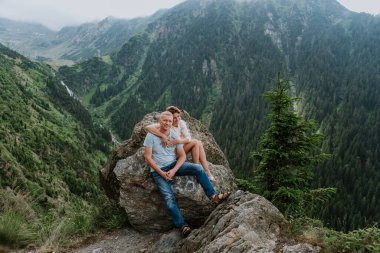 Çekici bir kadın ve kocası arka planda dağlarla birlikte kayaların üzerinde oturuyor. 