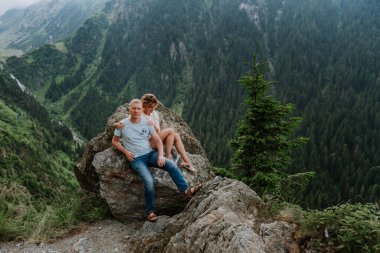 Çekici bir kadın ve kocası arka planda dağlarla birlikte kayaların üzerinde oturuyor. 