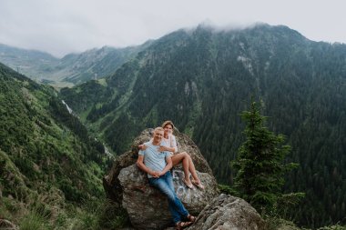 Çekici bir kadın ve kocası arka planda dağlarla birlikte kayaların üzerinde oturuyor. 