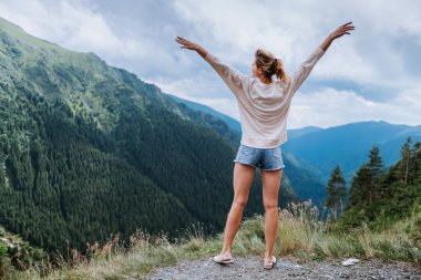 Genç bir kadın uçurumun kenarında duruyor ve elleri havada gökyüzüne bakıyor.