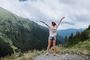 Genç bir kadın uçurumun kenarında duruyor ve elleri havada gökyüzüne bakıyor.