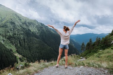 Genç bir kadın uçurumun kenarında duruyor ve elleri havada gökyüzüne bakıyor.