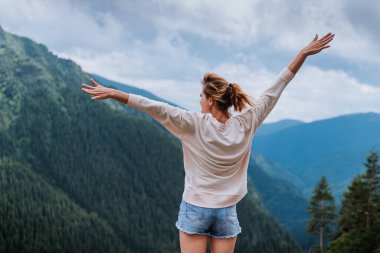 Genç bir kadın uçurumun kenarında duruyor ve elleri havada gökyüzüne bakıyor.