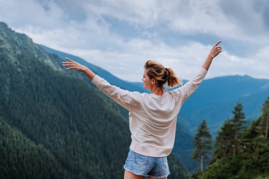 Genç bir kadın uçurumun kenarında duruyor ve elleri havada gökyüzüne bakıyor.