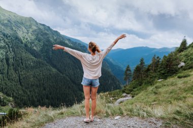 Genç bir kadın uçurumun kenarında duruyor ve elleri havada gökyüzüne bakıyor.