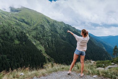 Genç bir kadın uçurumun kenarında duruyor ve elleri havada gökyüzüne bakıyor.