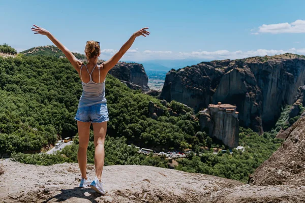 행복 한 젊은 여자는 손을 들고 카메라 앞에서 있다. 산과 함께 경치를 즐기는 여행자, 그리스의 메테오라 수도원 — 스톡 사진
