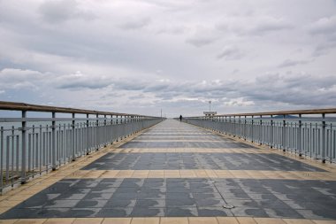 Doğrudan bir köprü sahili bulutlu gökyüzü ile deniz arasındaki boşluğa doğru terk eder. Köprü asfalt döşenmiş..