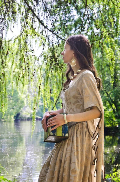 穿着旧衣服的年轻女子沿着公园的池塘岸边散步 背着一个老箱子 — 图库照片