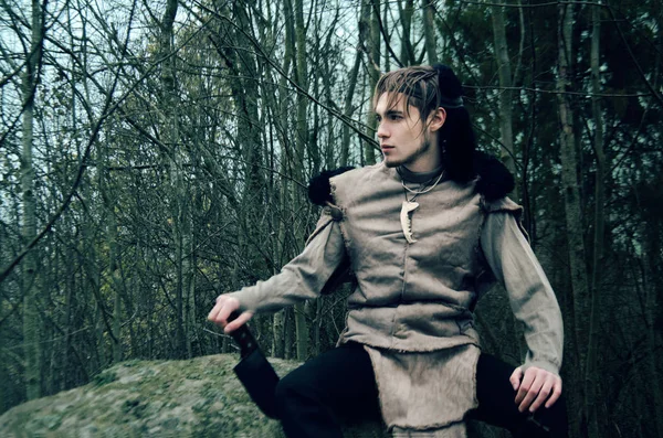 young man in the costume of a barbarian sitting on a stone in the dark forest