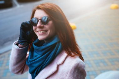 Kırmızı saçlı güzel kız tarafından bir pembe ceket ve bununla birlikte güneş gözlüklü mavi eşarp sokakta yürüyor.