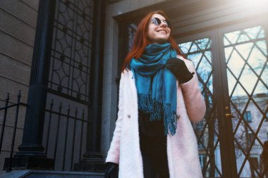 Kırmızı saçlı güzel kız tarafından bir pembe ceket ve bununla birlikte güneş gözlüklü mavi eşarp sokakta yürüyor.