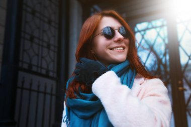 Kırmızı saçlı güzel kız tarafından bir pembe ceket ve bununla birlikte güneş gözlüklü mavi eşarp sokakta yürüyor.