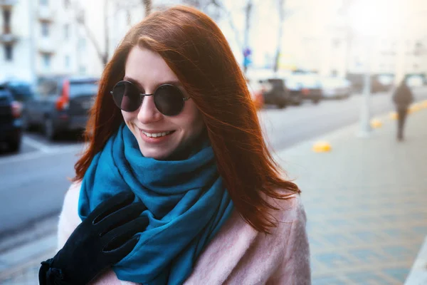 Belle fille aux cheveux rouges marche dans la rue avec un manteau rose et une écharpe bleue, avec des lunettes de soleil . — Photo