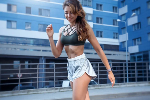 Chica deportiva de pelo largo en un elegante pantalones cortos de color plata y camiseta de color verde oscuro se está ejercitando sobre un fondo urbano.. La foto se hace durante el amanecer en una hermosa mañana de verano — Foto de Stock