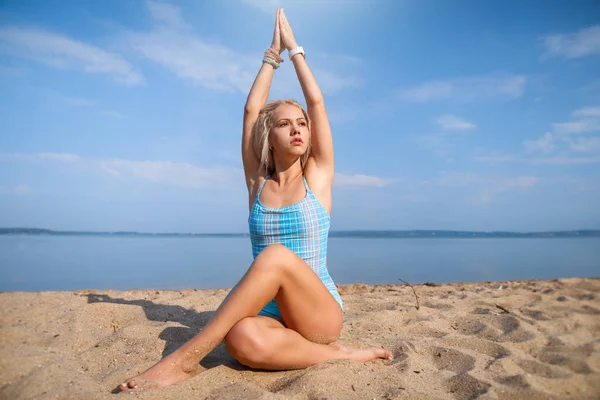 Блондинка длинноволосая девушка в синем костюме растягивается и занимается йогой на прекрасном пляже в солнечном свете восходящего солнца — стоковое фото