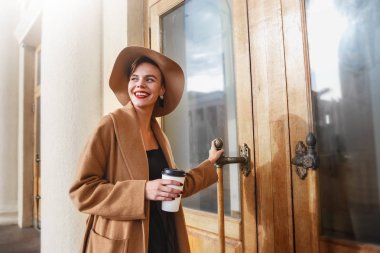 Kız kahverengi kabanlı bir kahverengi şapka yürüyüş ve şehir iç poz. Kız gülümsüyor, onun smartphone kontrol ve kahve içme kafe götürün.