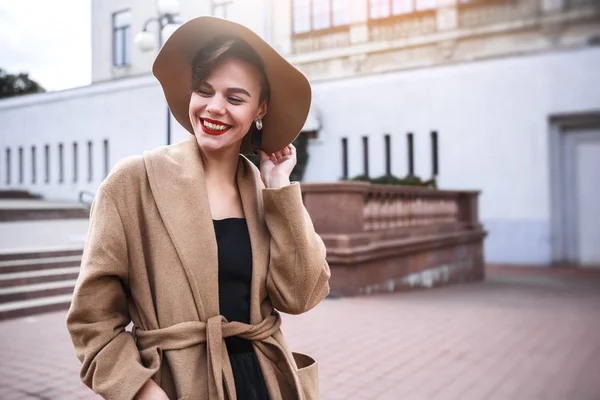 Chica con un abrigo marrón un sombrero marrón está caminando y posando en los interiores de la ciudad. La chica está sonriendo, revisando su teléfono inteligente y tomando café de la cafetería para llevar . — Foto de Stock