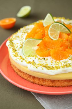 Turuncu bir tabakta, turuncu bir tabakta, yeşil gri bir zeminde, ezilmiş şam fıstığıyla süslenmiş, mandalina ve limon dilimleri ile süslenmiş, bisküvi soslu, limon kremalı turuncu meyveli kek..