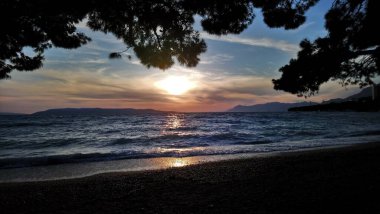 Hırvatistan. Makarska Riviera. Renkli günbatımının ve kumsaldan gökyüzünün muhteşem manzarası. Deniz kıyısı ve dalgalar. Kayalık plaj. Şehir parkı