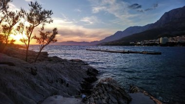 Hırvatistan. Makarska Riviera. Renkli günbatımının ve kumsaldan gökyüzünün muhteşem manzarası. Deniz kıyısı ve dalgalar. Kayalık plaj. Şehir parkı
