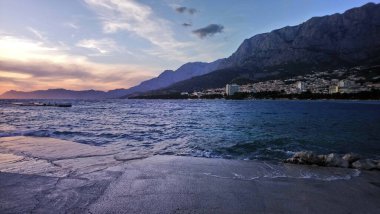 Hırvatistan. Makarska Riviera. Renkli günbatımının ve kumsaldan gökyüzünün muhteşem manzarası. Deniz kıyısı ve dalgalar. Kayalık plaj. Şehir parkı