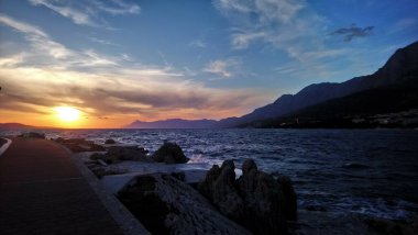 Hırvatistan. Makarska Riviera. Renkli günbatımının ve kumsaldan gökyüzünün muhteşem manzarası. Deniz kıyısı ve dalgalar. Kayalık plaj. Şehir parkı