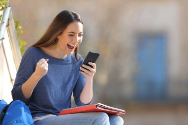 Heyecanlı öğrenci, üniversite kampüsünün dışında oturmuş akıllı telefon içeriğini kontrol ediyor.