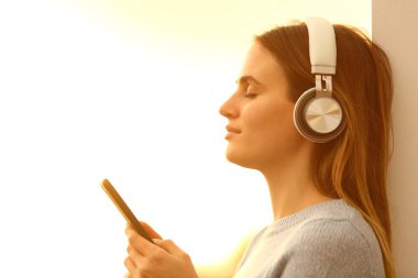 Kapalı gözlü, müzik dinleyen, kulaklık takan ve günbatımında akıllı bir telefon tutan rahat bir kızın portresi.