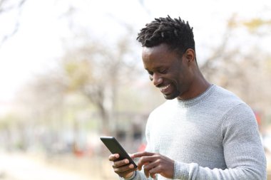 Güneşli bir günde parkta yürürken akıllı telefon kullanan mutlu siyah adam.
