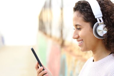 Müzik dinleyen, kulaklık takan ve sokakta akıllı telefonları kontrol eden mutlu bir Latin kızın yan görüntüsü.