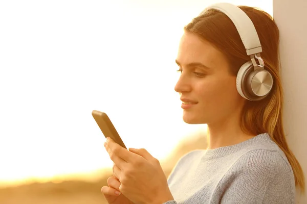 女孩戴着耳机听音乐 日落时分在海滩上看手机 — 图库照片