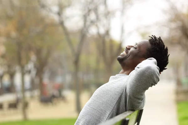 一个快乐的黑人坐在公园的长椅上悠闲自在的侧景画像 — 图库照片