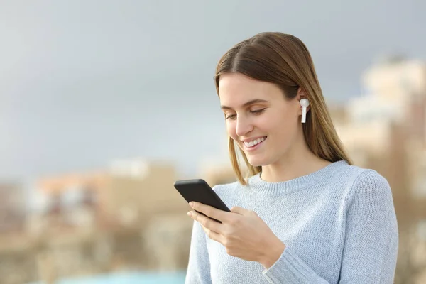 Happy teenage girl wearing wireless earbuds listening to music checking smart phone outside