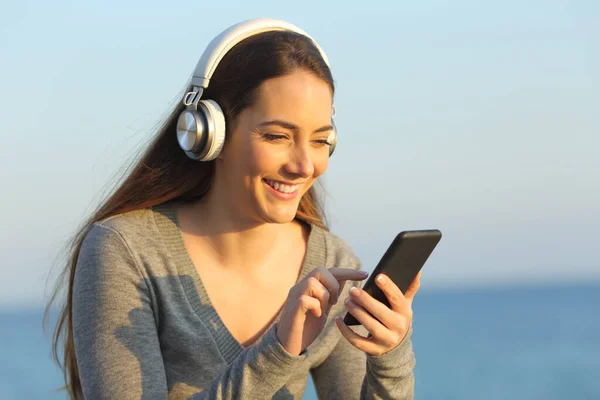 日没時にビーチに座っているスマートフォンを使用して音楽を聴く幸せな女性 — ストック写真