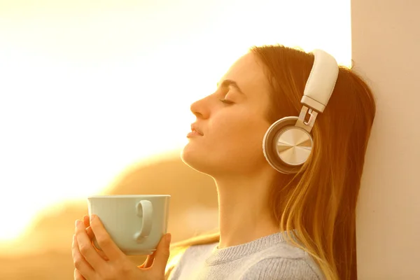 一个在日落时用耳机拿着咖啡杯听音乐的轻松女人的侧视图画像 — 图库照片