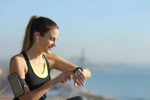 Happy runner wearing earbuds checking music on smartwatch outdoors in a city outskirts