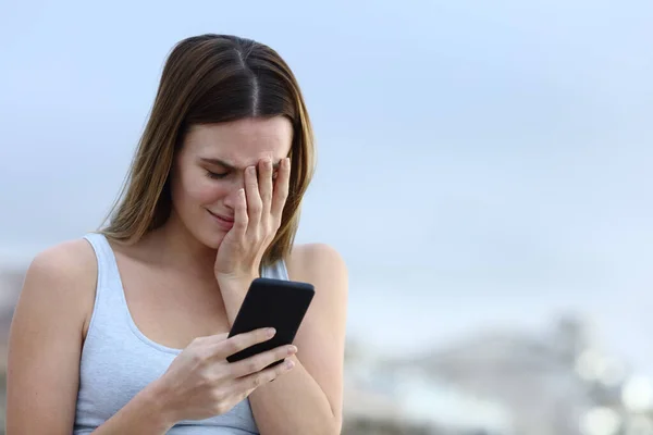 Sad woman crying checking bad news on mobile phone outdoors