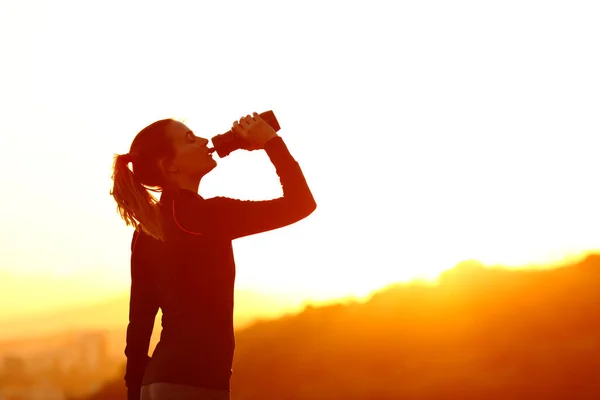 日落时用瓶子喝水的女跑步者的侧影 — 图库照片