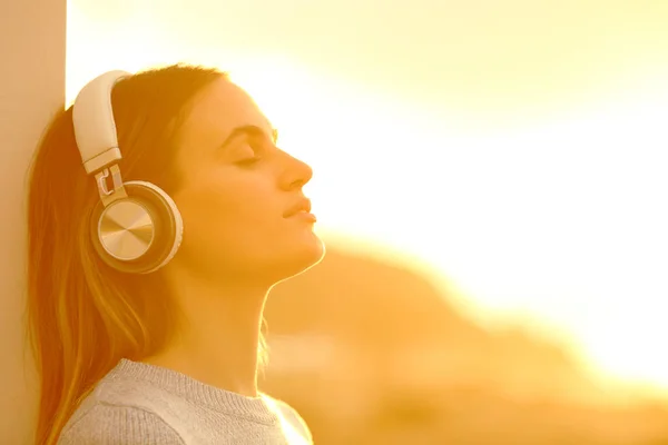 一名妇女在日落时穿着耳机听音乐 在海滩上休息的侧景肖像 — 图库照片