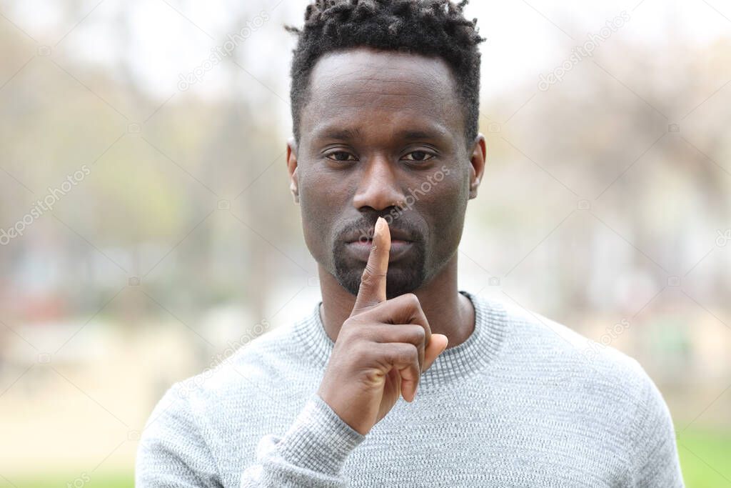 Front view portrait of a black man asking for silence with finger on lips standing in a park