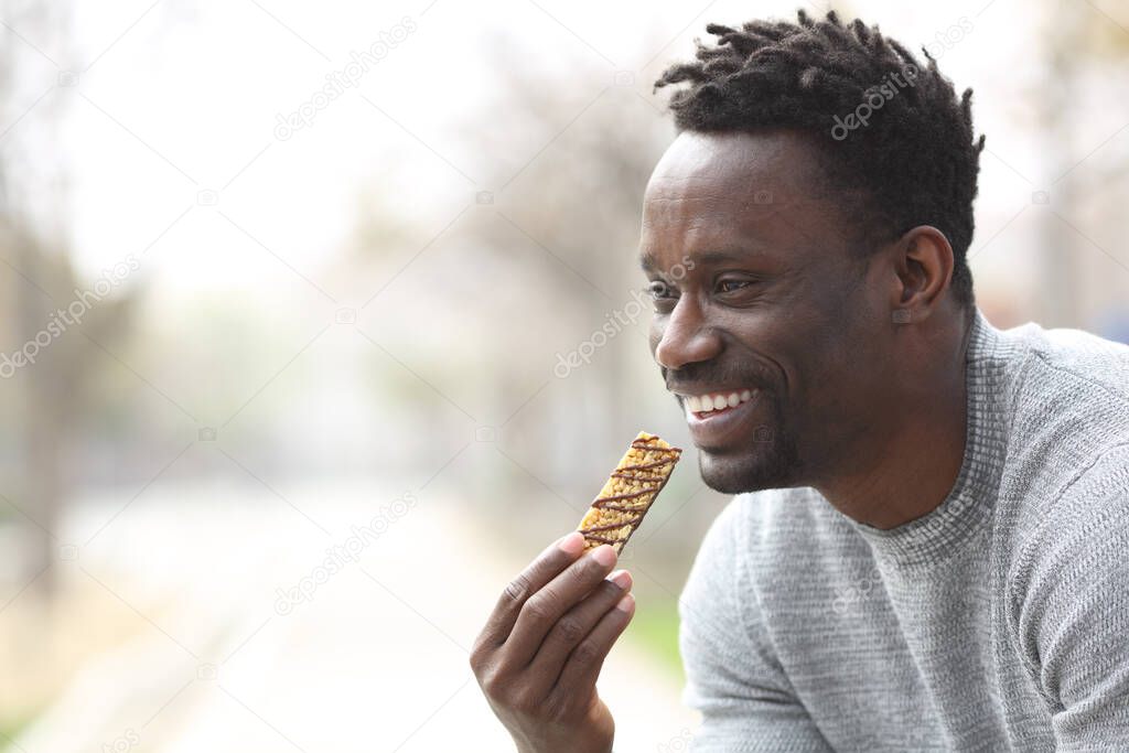 Happy black man eating a cereal bar in a park looking away