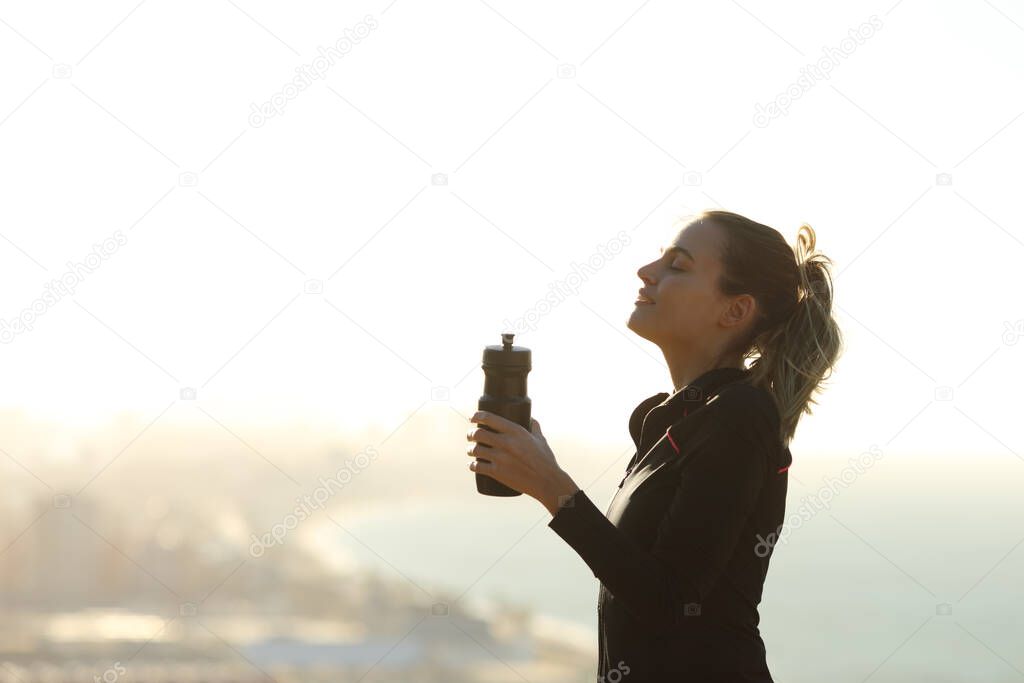 Profile of a runner woman relaxing breathing fresh air holding a water bottle with the city in the background