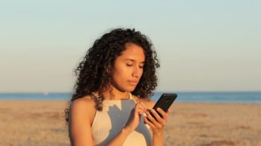 Heyecanlı Latin kadın cep telefonunu kontrol ediyor. Sahilde durmuş iyi haberler okuyor.